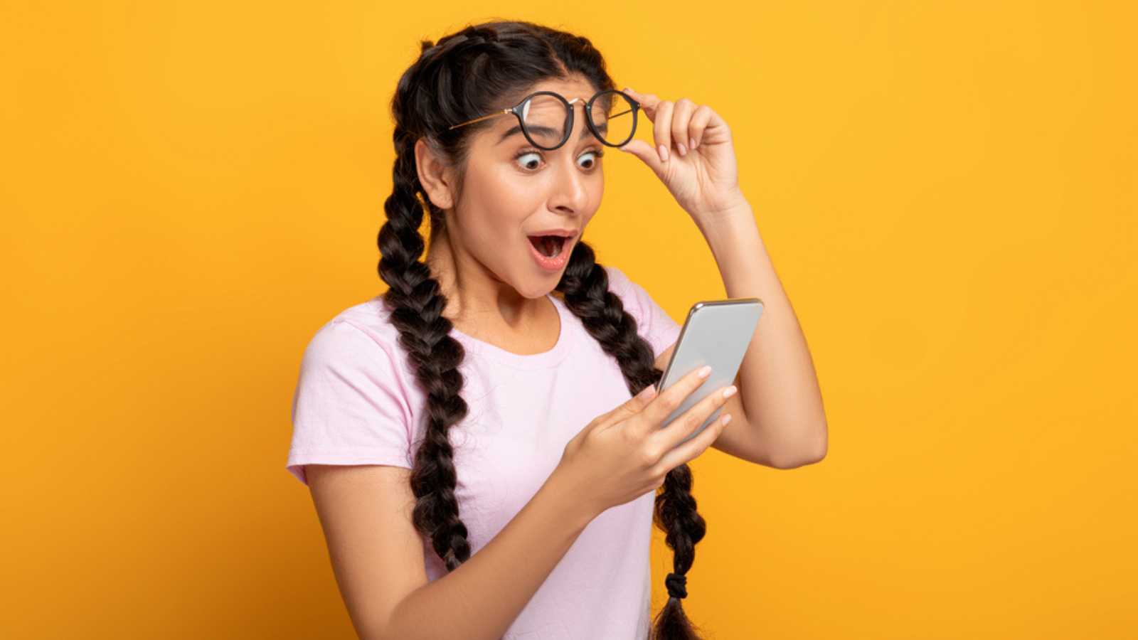 Wow, Great News. Portrait Of Excited Surprised Indian Woman Holding And Using Smartphone, Browsing Social Media. Emotional Young Lady Having Success, Taking Off Glasses, Yellow Studio Background